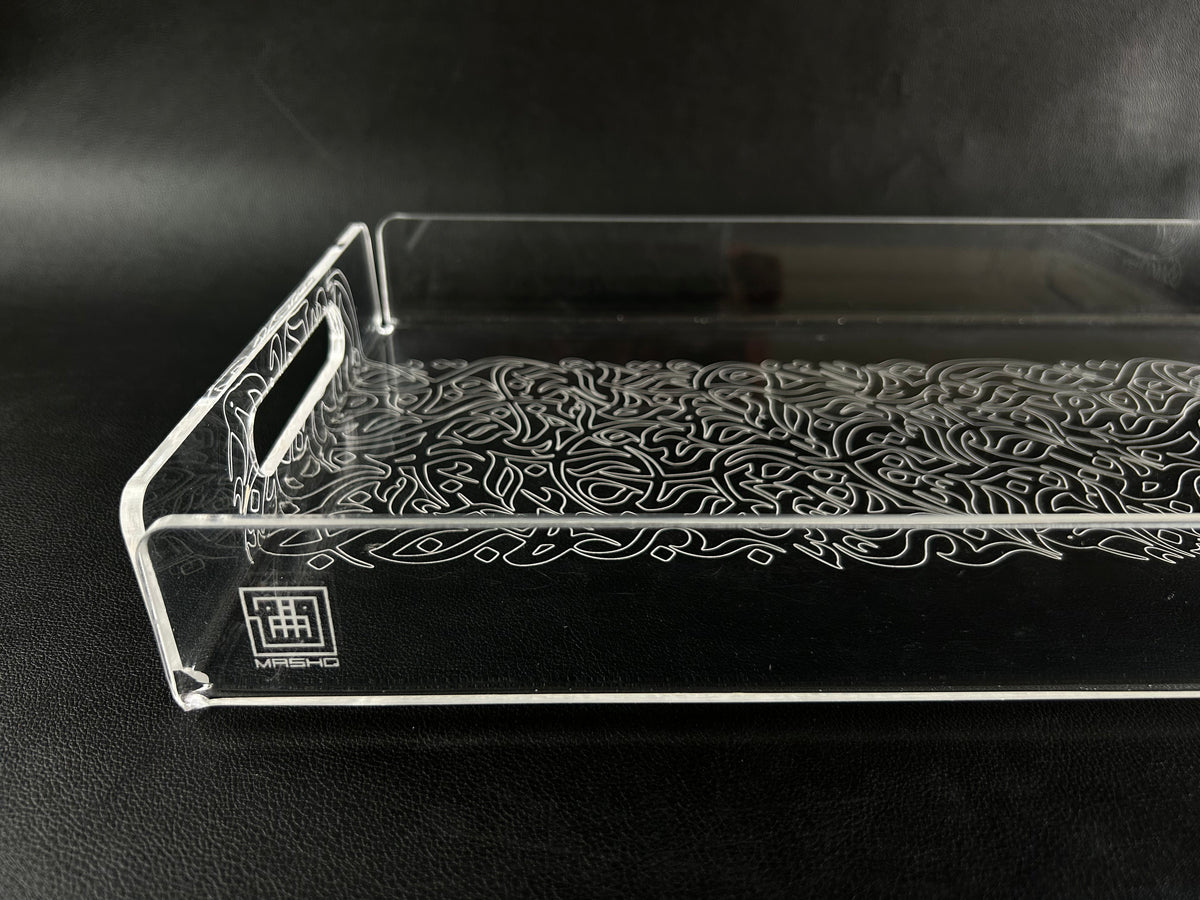 A clear acrylic serving tray engraved with Arabic calligraphy across its base, photographed on a dark surface; made in the UAE by MASHQ