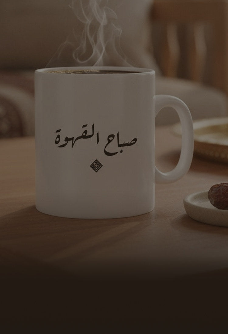 A white ceramic mug printed «صباح القهوة», steaming, beside a plate of dates on a coffee table
