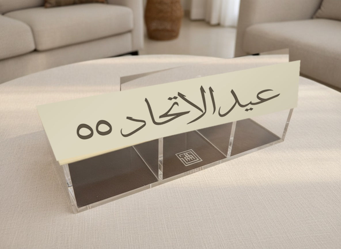 «عيد الاتحاد ٥٥» National Day dates box closed on a round majlis table, the Thuluth phrase cut through its cream lid, three clear compartments beneath
